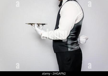 Profil Porträt von Butler oder Kellner in schwarzer Weste und weißen Handschuhen mit silbernem Serviertablett. Dienstleistungsbranche und professionelle Höflichkeit. Stockfoto