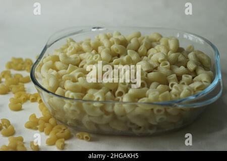 Gekochte Makkaroni-Pasta in einer quadratischen Glasschüssel. Aufgenommen auf weißem Hintergrund mit rohen Macaroni-Pastas Stockfoto