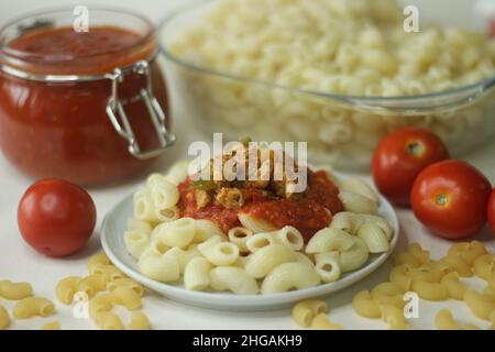 Makkaroni-Pasta mit roter Sauce und sautiertem Huhn und Paprika auf der Oberseite serviert. Shot mit gekochter Makkaroni-Pasta in einer Glasschüssel, rote Sauce in einem Glas Stockfoto