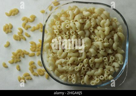 Gekochte Makkaroni-Pasta in einer quadratischen Glasschüssel. Aufgenommen auf weißem Hintergrund mit rohen Macaroni-Pastas Stockfoto
