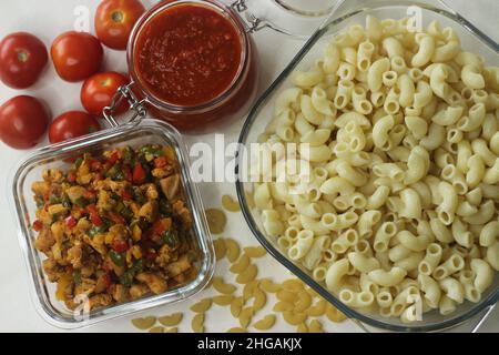 Gekochte Makkaroni-Pasta in einer quadratischen Glasschüssel. Shot auf weißem Hintergrund mit Tomatensoße in einem Glas, sautierten Huhn und Paprika und rohen ma Stockfoto