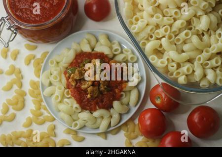 Makkaroni-Pasta mit roter Sauce und sautiertem Huhn und Paprika auf der Oberseite serviert. Shot mit gekochter Makkaroni-Pasta in einer Glasschüssel, rote Sauce in einem Glas Stockfoto