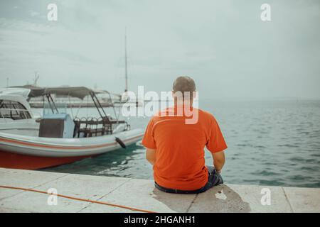 Ein Mann in einem roten T-Shirt sitzt am Meer vor dem Hintergrund seines Bootes. Fischer vor der Arbeit Stockfoto