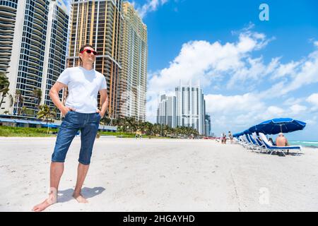 Ein junger Mann, der mit einer roten Sonnenbrille in Modekleidung am Strand in Sunny Isles, Miami, Florida, an der Küste steht, Skyline von Apartment con Stockfoto