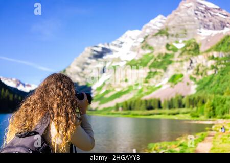 Rücken einer jungen Frau mit Kamera fotografiert Landschaft auf Aspen Maroon Bells felsigen Berggipfel mit Crater Lake Trail Wasser in Colorado im Sommer Stockfoto