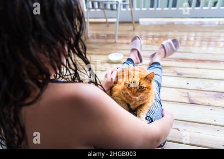 Junge Frau sitzt auf dem Boden mit Schoßkatze in den Armen streicheln streicheln streichelnd Katze orange Ingwer Kitty draußen zu Hause Haus Balkon Veranda Terrasse r Stockfoto