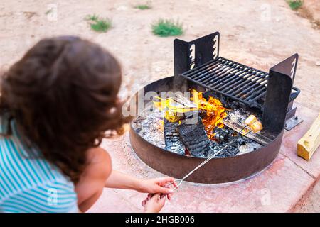 Frau, die beim Kochen von Sumpflaibchen am Spieß am Feuer auf dem Lagerfeuergrill mit roter Flamme im Hintergrund süßes Dessert grillt Stockfoto