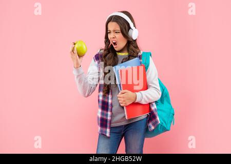 Staunendes Teenager-Mädchen mit Rucksack und Arbeitsbuch trägt Kopfhörer Blick auf Apfel, Mittagessen. Stockfoto