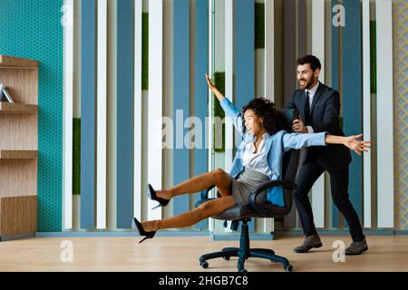 Zwei junge, fröhliche Geschäftsleute tragen Spaß, während sie auf Bürostühlen Rennen und lächeln - Kollegen feiern einen erfolgreichen Deal Stockfoto