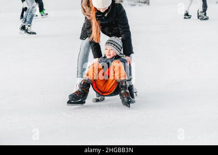 Weich, selektive Fokus.Mama mit Baby Junge 6 Jahre alt, lernen Zug, reiten Winterstadt Eislaufen. Das Kind steht auf, fällt auf Schlittschuhe, kniet, spielt Stockfoto