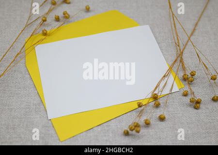 Modell mit Umschlag und leerer Karte. Ein gelber Umschlag auf weißem Stoff. Getrocknete Blüten (trockener Flachs) neben dem Umschlag. Minimalismus, Öko-Konzept. Stockfoto