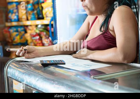 Besorgte Frau, die ihre Geschäftsstatistik-Tabelle überprüft. Schreiben mit einem umweltfreundlichen Pappstift und einem kleinen schwarzen Rechner auf einem großen Notizbuch. ba Stockfoto