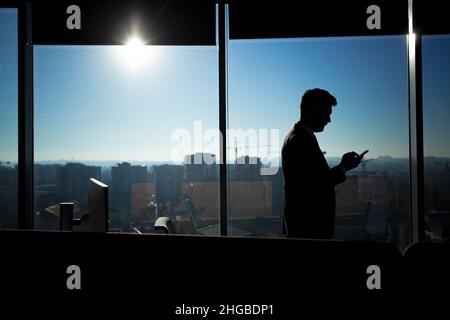 Silhouette des Geschäftsmannes im modernen Büro auf Fensterhintergrund, Mann nutzt Telefon Stockfoto