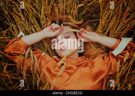 Ein Mädchen in einem goldenen Weizenfeld liegt in einem Kleid. Eine Frau schläft in der Natur, das Model hat die Augen geschlossen und genießt frische Luft Stockfoto