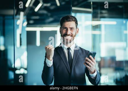 Glücklicher Geschäftsmann-Chef schaut auf die Kamera und lächelt, hält Telefon in den Händen, Mann gewinnt online und feiert den Sieg Stockfoto