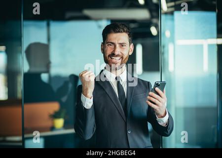 Glücklicher Geschäftsmann-Chef schaut auf die Kamera und lächelt, hält Telefon in den Händen, Mann gewinnt online und feiert den Sieg Stockfoto