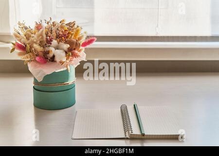 Offenes leeres Notizbuch mit türkisfarbenem Bleistift, schönes flauschiges Bouquet von getrockneten Blumen auf weißem Tisch. Stockfoto