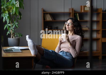 Junge, entspannte Mitarbeiterin, die sich am Arbeitsplatz mit einer Tasse Kaffee und Beinen auf dem Tisch im Büro ausruhte Stockfoto