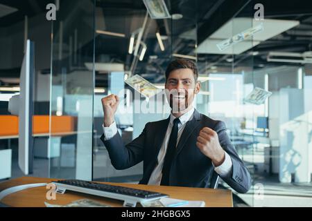 Schöner, fröhlicher junger Geschäftsmann, der an einem Schreibtisch sitzt, in einem modernen Büro arbeitet und Geld über den Kopf wirft. Glücklich lächelnder Mann wirft Geld in die A Stockfoto