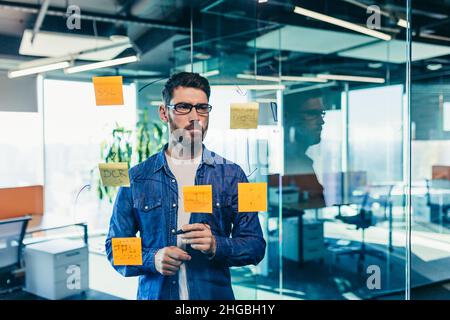 Fokussierter und denkender Geschäftsmann, der auf bunten Notizen an einer Glaswand schreibt, erstellt der Unternehmer eine visuelle To-Do-Liste, Geschäftsziele für Stockfoto