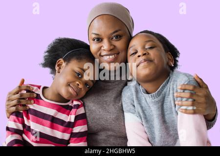 Porträt der fürsorglichen islamischen afrikanischen Mutter in Hijab umarmt ihre zwei niedlichen Töchter auf lila Hintergrund. Konzept der Familie, Liebe und Lebensstile. Stockfoto