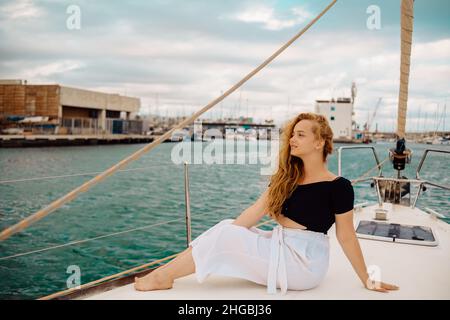 Ein wunderschönes langhaariges Mädchen sitzt auf dem Bug einer versperrten Yacht und genießt das Leben. Die Blondine ruht im offenen Meer oder Meer. Stockfoto