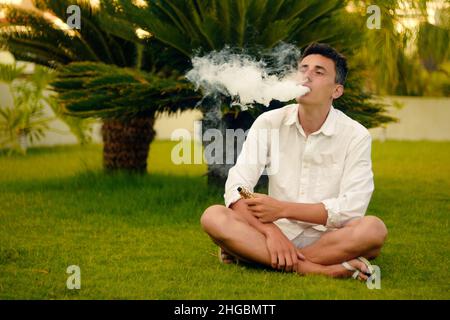 Ein Vape-Mann im Urlaub, unter einer Palme. Ruhe, Rauchen, Nikotin, Genuss, Gefahr. Flüssigkeit abdampfen. Dampfende THC e Zigarette. Stockfoto