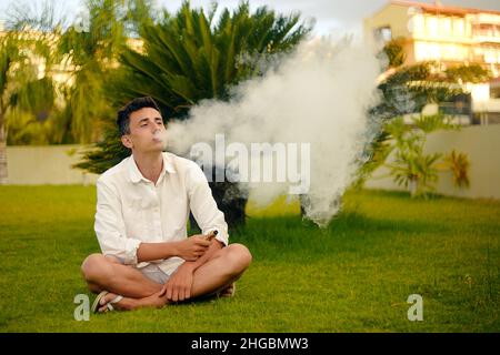 Ein Vape-Mann im Urlaub, unter einer Palme. Ruhe, Rauchen, Nikotin, Genuss, Gefahr. Flüssigkeit abdampfen. Dampfende THC e Zigarette. Stockfoto