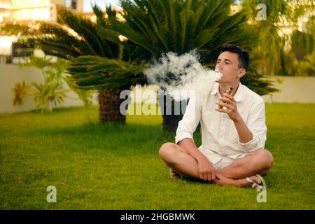 Ein Vape-Mann im Urlaub, unter einer Palme. Ruhe, Rauchen, Nikotin, Genuss, Gefahr. Flüssigkeit abdampfen. Dampfende THC e Zigarette. Stockfoto