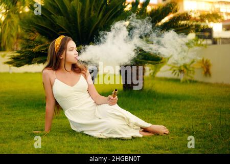 Eine Vape-Frau im Urlaub, unter einer Palme. Ruhe, Rauchen, Nikotin, Genuss, Gefahr. Flüssigkeit abdampfen. Dampfende THC e Zigarette. Stockfoto