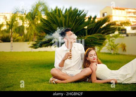 Ein Mann und eine Frau. Pärchen im Urlaub, unter einer Palme. Ruhe, Rauchen, Nikotin, Genuss, Gefahr. Flüssigkeit abdampfen. Dampfende THC e Zigarette. Stockfoto