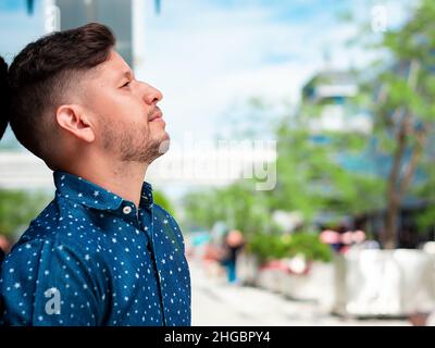 Ein junger, hübscher hispanischer Latino-Mann mit Bart und kurzen Haaren, in einem blauen Hemd gekleidet, posiert im Profil und lehnt sich mit dem sk an eine Glaswand Stockfoto