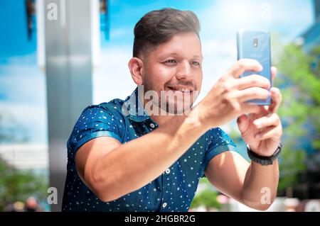 Der junge, gutaussehende lateinamerikanische Junge mit Bart und kurzen Haaren, gekleidet in ein blaues Hemd, ist glücklich und lächelt und macht ein Foto mit seinem Handy in einer Ur Stockfoto