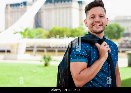 Junger, attraktiver lateinamerikanischer Junge, mit Bart und kurzen Haaren, in einem blauen Kurzarmhemd gekleidet, mit einer schwarzen Jacke über der Schulter, aufgesetzt Stockfoto