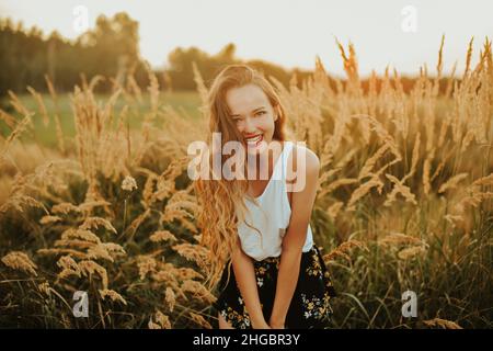 Wunderschöne Frau in einem Weizenfeld auf einem Sonnenuntergang Hintergrund. Ein modisches Mädchen mit langen Haaren freut sich, lacht, genießt Leben und Sommer, Natur, Glück. Stockfoto