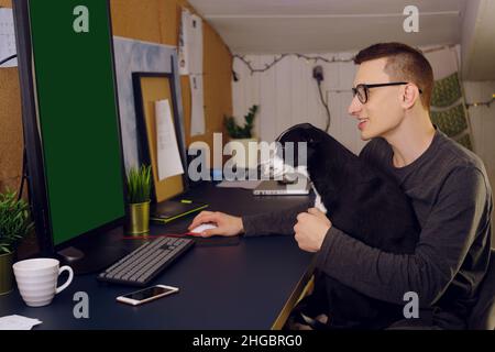 Bleiben Sie zu Hause, ein Mann sitzt zu Hause an einem Computer, ein Hund stört die Arbeit. Ein Mann arbeitet zu Hause, der Hund sitzt in seinen Armen und schaut auf den Computer. C Stockfoto