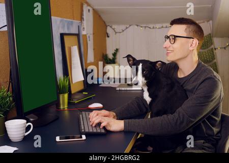 Bleiben Sie zu Hause, ein Mann sitzt zu Hause an einem Computer, ein Hund stört die Arbeit. Ein Mann arbeitet zu Hause, der Hund sitzt in seinen Armen und schaut auf den Computer. C Stockfoto