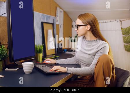 Bleiben Sie zu Hause, ein schickes Mädchen arbeitet in einer heimeligen Atmosphäre. Mädchen Freiberuflerin arbeitet an einem Computer mit Brille, Blick auf einen grünen Bildschirm auf einem Computer. Der Stockfoto