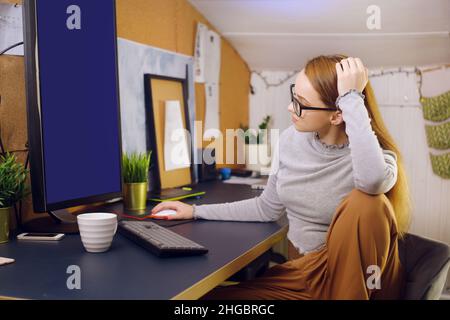 Bleiben Sie zu Hause, ein schickes Mädchen arbeitet in einer heimeligen Atmosphäre. Mädchen Freiberuflerin arbeitet an einem Computer mit Brille, Blick auf einen grünen Bildschirm auf einem Computer. Der Stockfoto