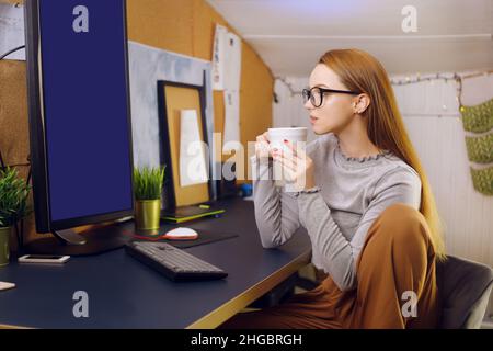 Bleiben Sie zu Hause, ein schickes Mädchen arbeitet in einer heimeligen Atmosphäre. Mädchen Freiberuflerin arbeitet an einem Computer mit Brille, Blick auf einen grünen Bildschirm auf einem Computer. Der Stockfoto