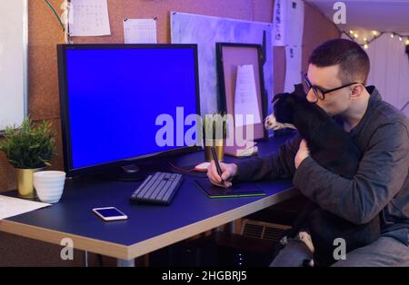 Der Hund sitzt in den Händen des Mannes. Ein Mann arbeitet zu Hause, ein Grafiker, ein Schöpfer arbeitet zu Hause auf freiberuflich mit einem Hund. Das Haustier macht es schwierig Stockfoto