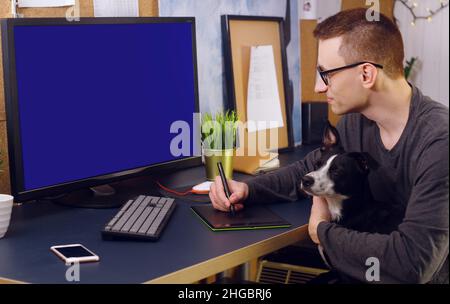 Der Hund sitzt in den Händen des Mannes. Ein Mann arbeitet zu Hause, ein Grafiker, ein Schöpfer arbeitet zu Hause auf freiberuflich mit einem Hund. Das Haustier macht es schwierig Stockfoto