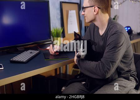 Der Hund sitzt in den Händen des Mannes. Ein Mann arbeitet zu Hause, ein Grafiker, ein Schöpfer arbeitet zu Hause auf freiberuflich mit einem Hund. Das Haustier macht es schwierig Stockfoto