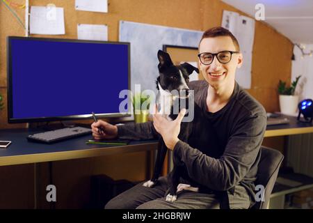 Der Hund sitzt in den Händen des Mannes. Ein Mann arbeitet zu Hause, ein Grafiker, ein Schöpfer arbeitet zu Hause auf freiberuflich mit einem Hund. Das Haustier macht es schwierig Stockfoto