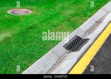 Graue Rinne eines Regenwasserentwässerungssystems auf der Seite der Asphaltstraße mit gelben Markierungen und Kanalisation Klärgrube in grünem Rasen, Betonentwässerung di Stockfoto