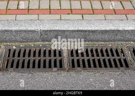 Auffangbecken Gitter des Gitters des Abflusssystems für die Ableitung von Regenwasser von der Straßenoberfläche mit einer asphaltierten Straße an der Bordsteinkante mit einem Steinziegel Stockfoto