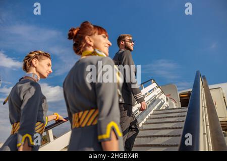Lächelnder Flugkapitän und zwei Stewardessen-Frauen, die unter blauem Himmel eine Flugzeugtreppe besteigen Stockfoto
