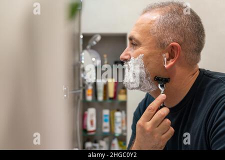 Ein hübscher weißhaariger bärtiger Mann, der sich seinen Bart abrasieren und in einen Spiegel schaut. Konzept eines sich ändernden Stils Stockfoto