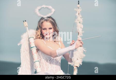 Unschuldiges Mädchen mit Engelsflügeln, das mit Pfeil und Bogen gegen den blauen Himmel und weiße Wolken steht. Valentinstag. Kleines Engelmädchen gegen sonnigen Himmel Stockfoto