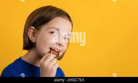 Kleines Mädchen 8-10 Jahre alt in Basic Blue T-Shirt beißen Golden Physical Bitcoin auf gelbem Hintergrund. Kinder Finanzielle Kompetenz. Kryptowährung und Blockchain-Konzept. Stockfoto